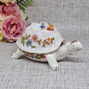 May include: A white ceramic turtle ornament with a floral pattern on its shell. The shell is decorated with colourful flowers and butterflies. The turtle's legs and head are white with gold detailing. A decorative item.