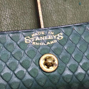 Pu&ograve; includere: Primo piano di un articolo in pelle verde con motivo a diamante. Il testo "MADE IN STANLEYS ENGLAND" &egrave; impresso in oro. &Egrave; visibile un bottone automatico in metallo dorato.
