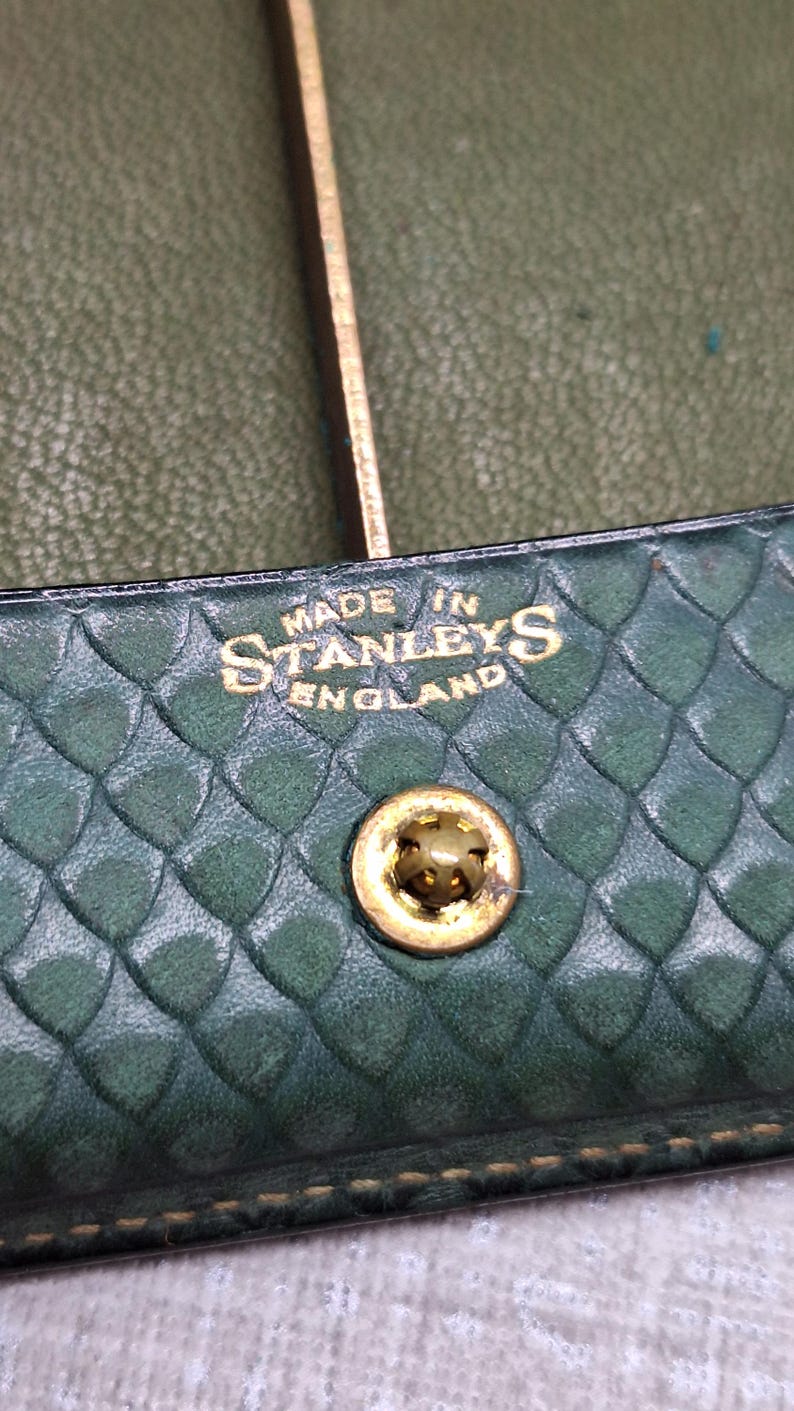 Pu&ograve; includere: Primo piano di un articolo in pelle verde con motivo a diamante. Il testo "MADE IN STANLEYS ENGLAND" &egrave; impresso in oro. &Egrave; visibile un bottone automatico in metallo dorato.