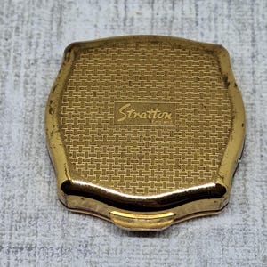 May include: A vintage gold-toned Stratton compact with a textured, woven pattern on the lid. The compact features the brand name "Stratton England" in a cursive font. The compact has a rectangular shape with rounded edges.