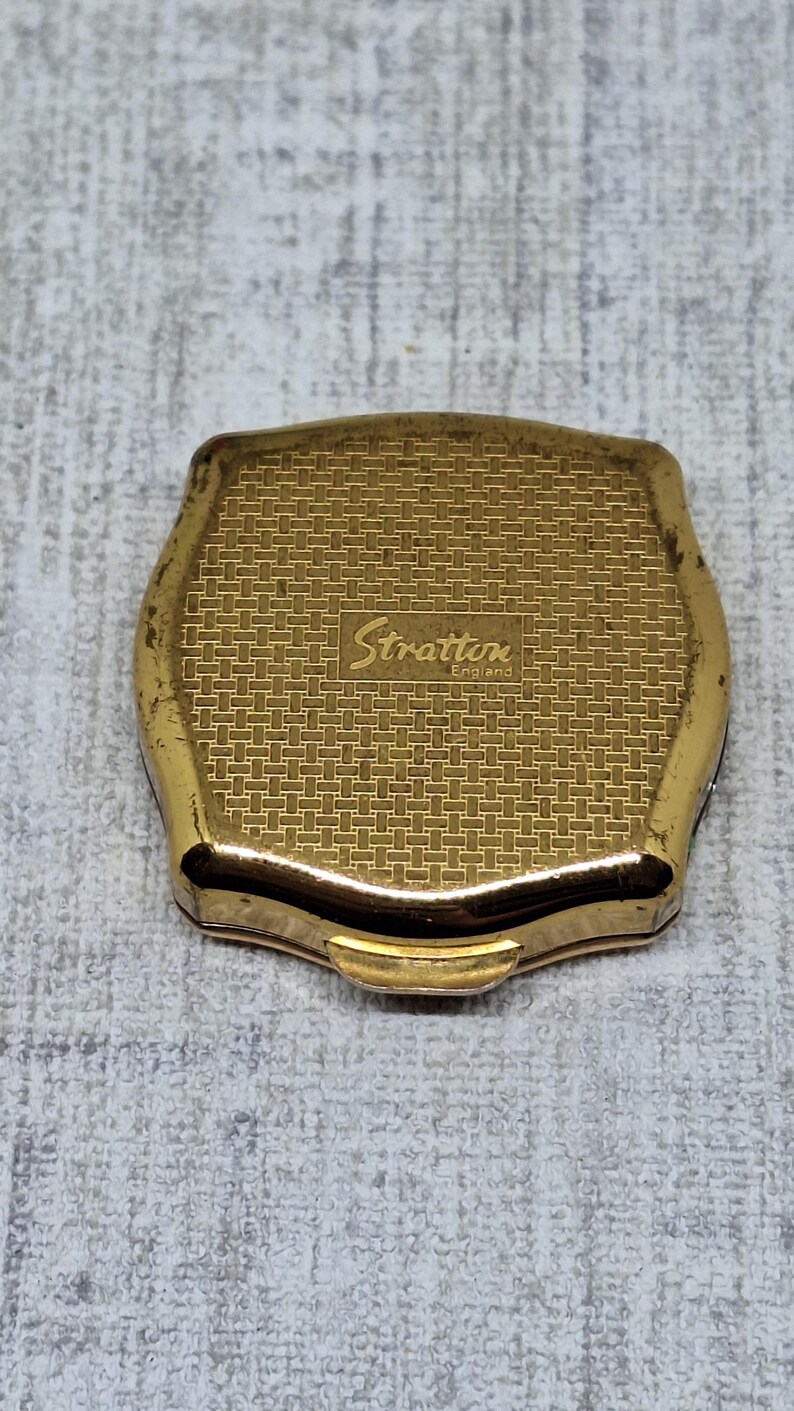 May include: A vintage gold-toned Stratton compact with a textured, woven pattern on the lid. The compact features the brand name "Stratton England" in a cursive font. The compact has a rectangular shape with rounded edges.