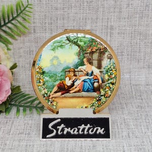 Op de afbeelding: Een vintage Stratton compacte spiegel met een goudkleurig frame. De compact heeft een geschilderde scène van een stel in een tuin. Het woord "Stratton" wordt weergegeven op een zwart-wit label. De compact is rond en heeft een scharnierende opening.