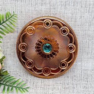 Puede incluir: Un compacto redondo de color dorado con un diseño decorativo que presenta una joya central de color turquesa. El compacto tiene un borde circular elevado y un patrón repetitivo de círculos y motivos florales.