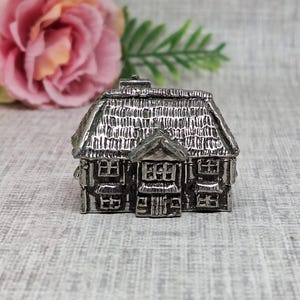 Könnte beinhalten: Ein detailliertes, silberfarbenes Miniaturhaus mit Strohdach und mehreren Fenstern. Das kleine Haus ist ein dekorativer Gegenstand, möglicherweise ein Sammlerstück oder eine Schmuckschatulle. Das Haus ist aus Metall gefertigt und weist aufwendige Details auf.