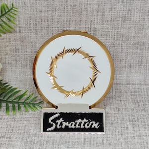 May include: A white compact mirror with a gold trim and a gold lightning bolt design. The mirror is closed and has a black label with the word "Stratton" on it.