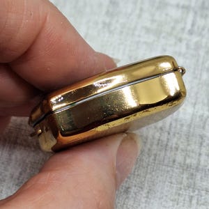 May include: A small, rectangular, gold-colored metal box with a hinged lid. The box has a shiny, reflective surface and appears to be empty. The box is held in a person's hand.