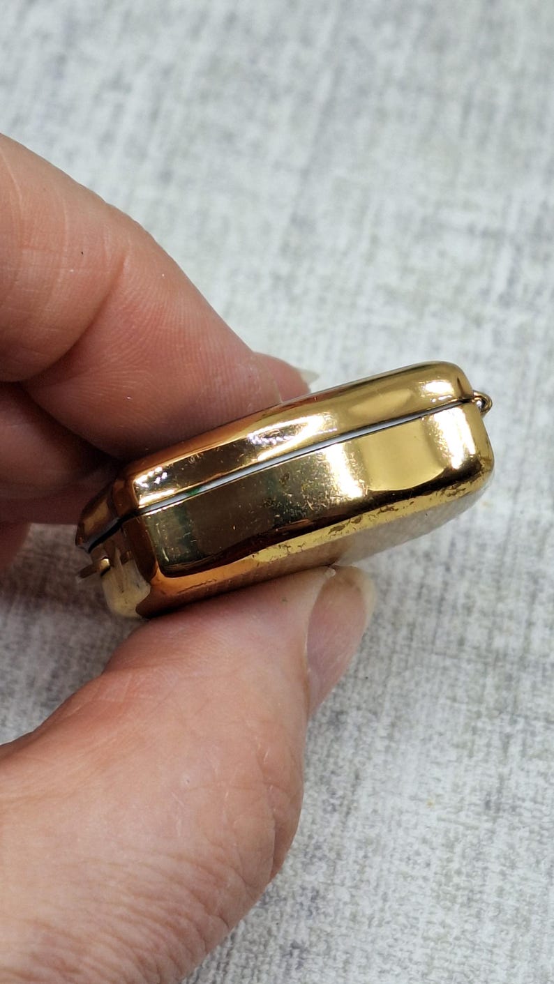 May include: A small, rectangular, gold-colored metal box with a hinged lid. The box has a shiny, reflective surface and appears to be empty. The box is held in a person's hand.