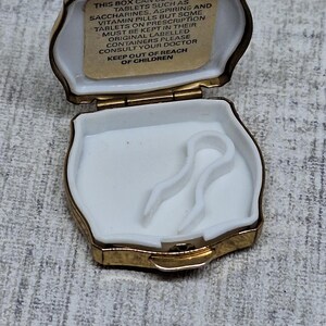 May include: Open, gold-trimmed pill box with a white interior. The box has a hinged lid and a small compartment with a white plastic insert. A label inside the lid provides instructions. The box is designed to hold tablets.