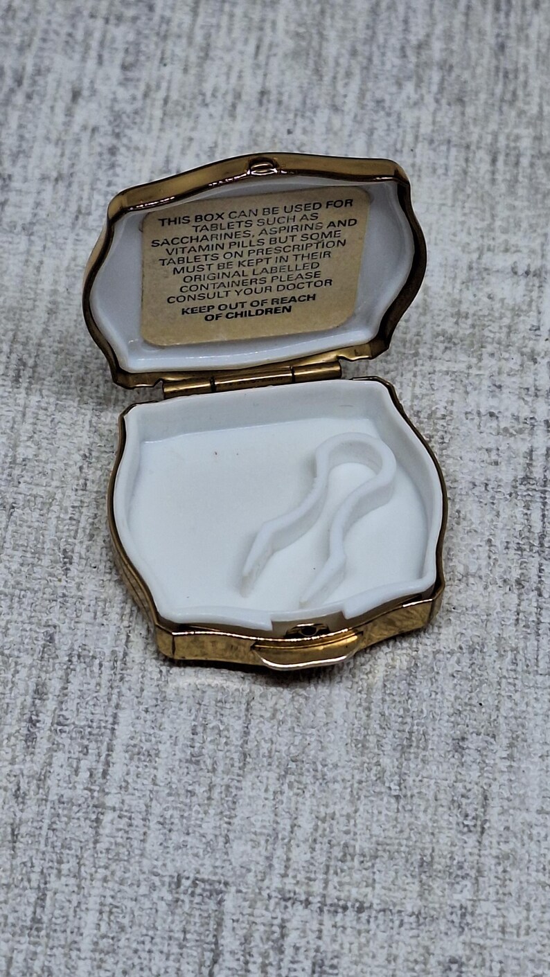 May include: Open, gold-trimmed pill box with a white interior. The box has a hinged lid and a small compartment with a white plastic insert. A label inside the lid provides instructions. The box is designed to hold tablets.