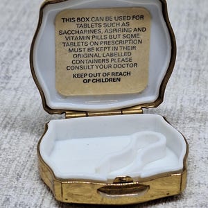 May include: Open, gold-toned pill box with a white interior. The box has a hinged lid and a rectangular shape with rounded edges. Text inside the lid reads: "THIS BOX CAN BE USED FOR TABLETS...KEEP OUT OF REACH OF CHILDREN."