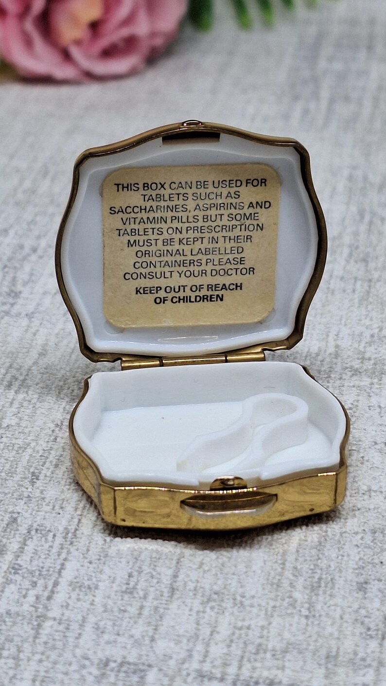 May include: Open, gold-toned pill box with a white interior. The box has a hinged lid and a rectangular shape with rounded edges. Text inside the lid reads: "THIS BOX CAN BE USED FOR TABLETS...KEEP OUT OF REACH OF CHILDREN."