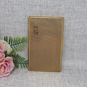 Peut inclure: Un étui à cigarettes doré de forme rectangulaire avec un motif texturé en forme de grille. Un petit emblème avec des initiales stylisées est centré sur le devant. L'étui est fermé. Fond en tissu gris clair.