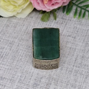 以下が含まれることがあります： 長方形の緑色の石が、銀色の金属ケースにセットされています。金属ケースのベースには、装飾的な彫刻デザインが施されています。オブジェクトは、ニュートラルなパターン化された背景に配置され、背景には花があります。