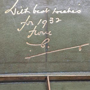 Pu&ograve; includere: Primo piano di un oggetto in pelle verde vintage con scritte dorate. Il testo recita "With best wishes for 1932 from B." Sono visibili anche una cornice in metallo dorato e un elemento di fissaggio metallico circolare.