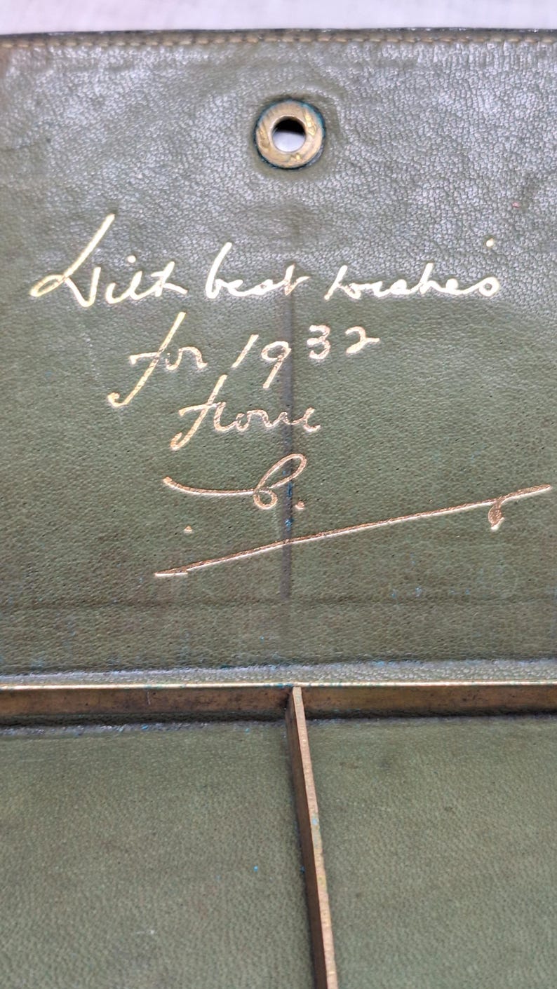 Pu&ograve; includere: Primo piano di un oggetto in pelle verde vintage con scritte dorate. Il testo recita "With best wishes for 1932 from B." Sono visibili anche una cornice in metallo dorato e un elemento di fissaggio metallico circolare.