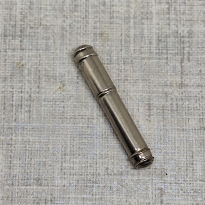 Puede incluir: Un objeto cilíndrico de color plateado con una parte superior e inferior redondeadas. El objeto parece ser de metal y tiene un diseño segmentado, con una bisagra o junta central. El artículo está sobre una superficie texturizada de color gris claro.