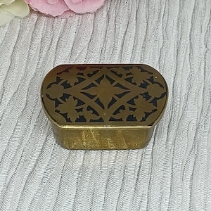 以下が含まれることがあります： 黒い内側を持つ、小さな楕円形の金色の金属製の宝箱。蓋には、スタイリッシュな葉と花がカットアウトされた装飾的なデザインが施されています。