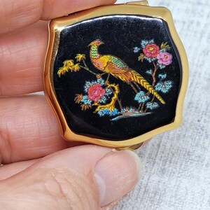 May include: A small, vintage compact with a gold-tone frame. The black enamel top features a colorful illustration of a pheasant perched on a branch with pink and blue flowers. The compact is held in a hand.