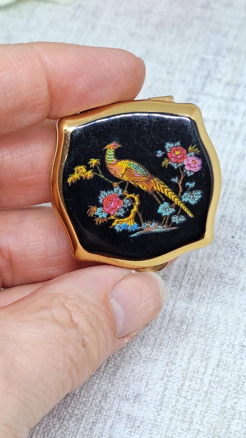 May include: A small, vintage compact with a gold-tone frame. The black enamel top features a colorful illustration of a pheasant perched on a branch with pink and blue flowers. The compact is held in a hand.