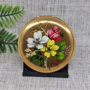 以下が含まれることがあります： 丸い金色のコンパクトミラー。ピンクのバラ、白と黄色の花、緑の葉のフローラルデザイン。コンパクトは、ニュートラルな背景に対して黒いスタンドに表示されています。