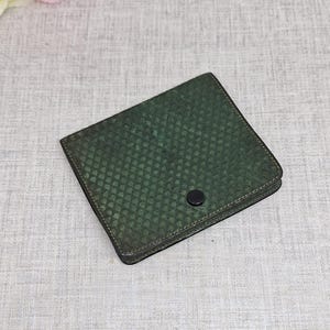 Pu&ograve; includere: Un portafoglio quadrato verde scuro con un motivo strutturato a forma di scaglia. Il portafoglio ha una chiusura a scatto nera e un bordo nero. Il portafoglio &egrave; probabilmente in pelle o in un materiale simile.