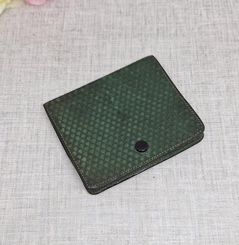 Pu&ograve; includere: Un portafoglio quadrato verde scuro con un motivo strutturato a forma di scaglia. Il portafoglio ha una chiusura a scatto nera e un bordo nero. Il portafoglio &egrave; probabilmente in pelle o in un materiale simile.