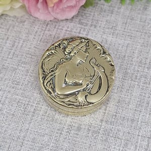 May include: A circular, antique brass trinket box with an Art Nouveau design. The lid has a relief of a woman playing a stringed instrument, surrounded by flowing lines and floral elements. The box is set against a textured grey background.