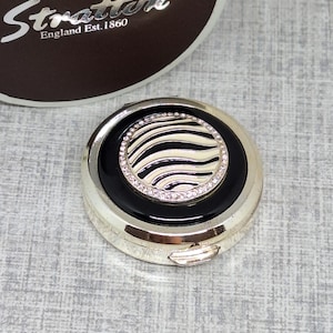 May include: A circular, silver-coloured compact featuring a black and white zebra-striped central design. The centre is encircled by a black band and a ring of clear rhinestones. The compact has a hinged opening, with the brand name "Stratton England Est. 1860" visible.