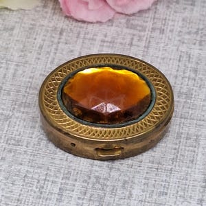 May include: A vintage, round, brass compact with an amber-colored, faceted stone center. The compact has a decorative, etched border and a small clasp. The stone is set in a teal-colored frame.