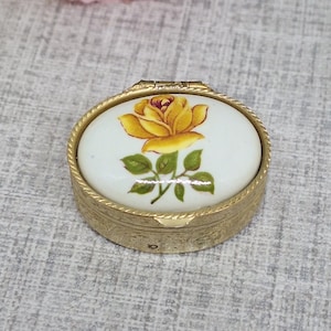 Może przedstawiać: Małe, owalne pudełko na drobiazgi z otwieraną pokrywką. Pokrywka przedstawia malowaną żółtą różę z zielonymi liśćmi na białym tle. Pudełko ma złotą metalową ramę z wzorem w kształcie liny na krawędziach. Około 5 cm długości.