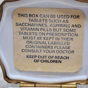 May include: Open, white, octagonal pill box with a gold-colored trim. The interior features a beige label with black text advising on the use of the box for tablets and to keep out of reach of children.