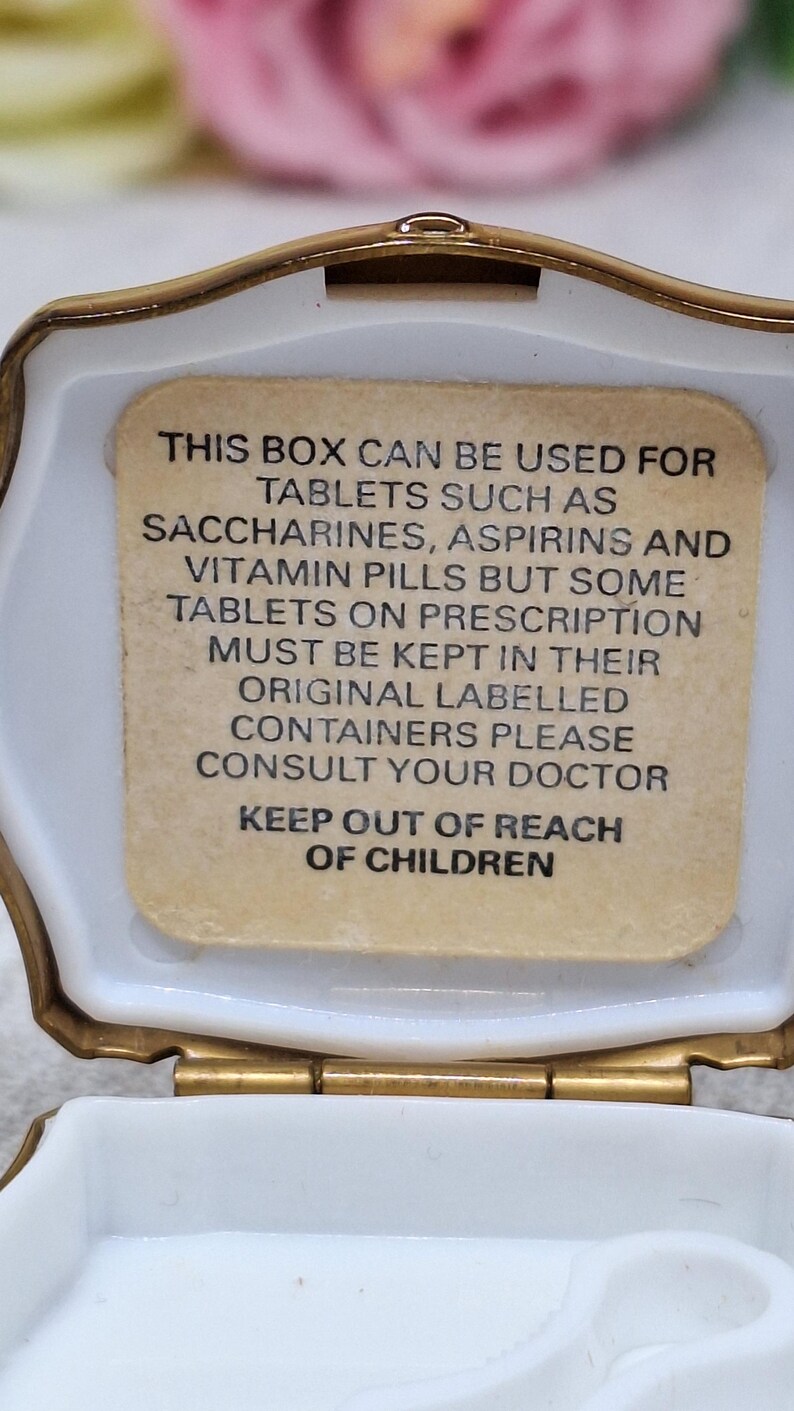 May include: Open, white, octagonal pill box with a gold-colored trim. The interior features a beige label with black text advising on the use of the box for tablets and to keep out of reach of children.