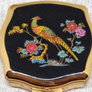 May include: Vintage compact mirror with a gold-tone frame and a black enamel top. The top features a colorful illustration of a pheasant perched on a flowering branch with red and pink blossoms. A classic vanity accessory.