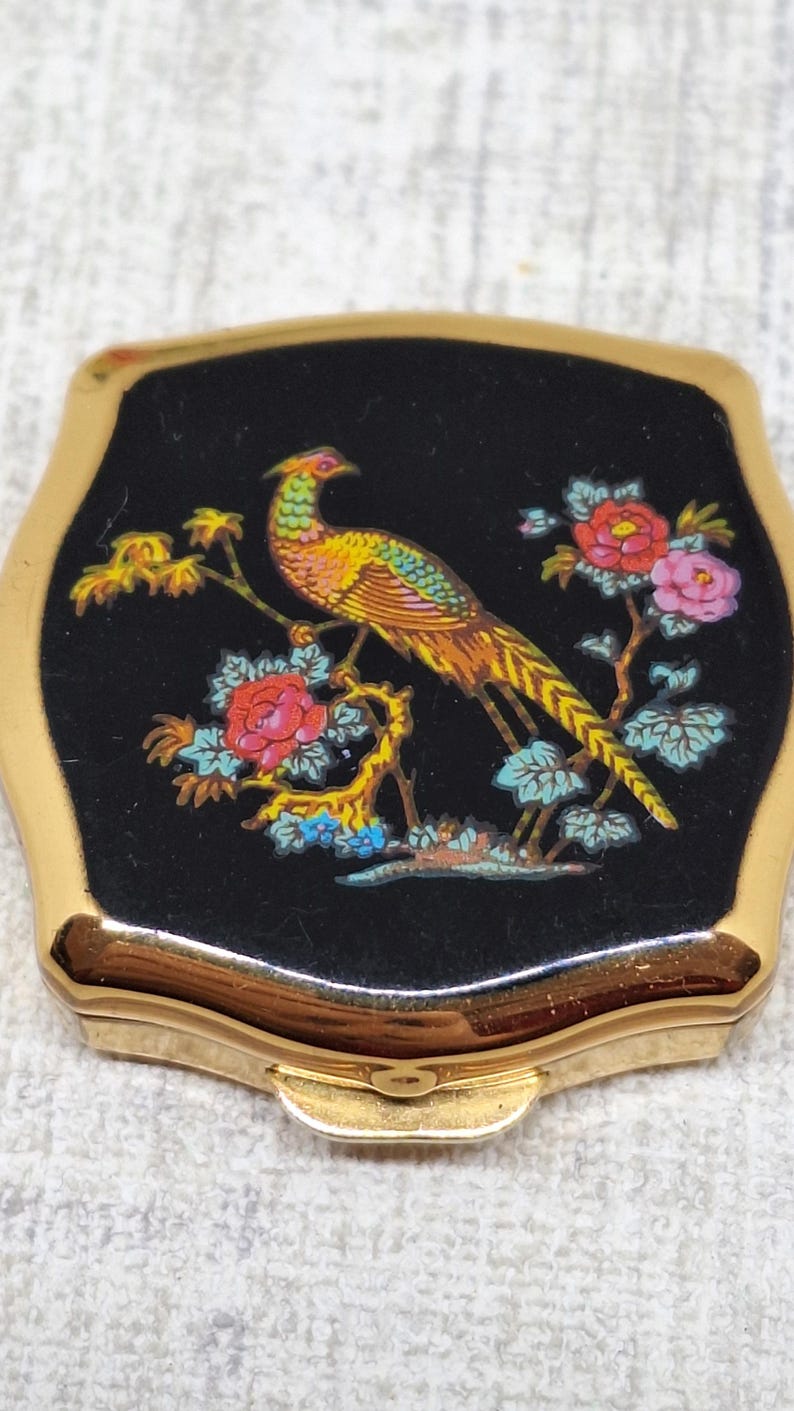 May include: Vintage compact mirror with a gold-tone frame and a black enamel top. The top features a colorful illustration of a pheasant perched on a flowering branch with red and pink blossoms. A classic vanity accessory.