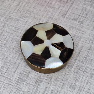 May include: A circular decorative item with a soccer ball design. The top surface has a pattern of dark brown, white, and cream-coloured segments. It has a brass-coloured metal rim. Likely a decorative box or small container.
