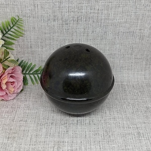Könnte beinhalten: Dunkelgrünes, kugelförmiges Dekorationsobjekt mit drei kleinen Löchern oben. Das Objekt hat eine glänzende Oberfläche und einen abgerundeten Boden. Es ist wahrscheinlich eine dekorative Schale oder ein Behälter.