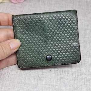 Pu&ograve; includere: Un piccolo portafoglio quadrato verde scuro con una superficie strutturata a motivo diamantato. Il portafoglio ha una chiusura a scatto nera e sembra essere in pelle. I bordi sono cuciti con un filo pi&ugrave; scuro.