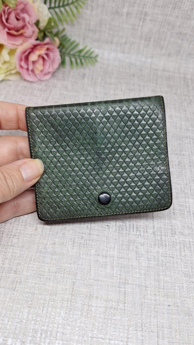 Pu&ograve; includere: Un piccolo portafoglio quadrato verde scuro con una superficie strutturata a motivo diamantato. Il portafoglio ha una chiusura a scatto nera e sembra essere in pelle. I bordi sono cuciti con un filo pi&ugrave; scuro.