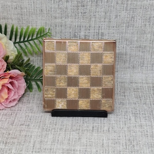 May include: A vintage, square compact mirror with a gold-toned frame and a checkerboard pattern in shades of brown and gold. The compact is displayed on a black stand, with pink and white flowers and green foliage in the background.