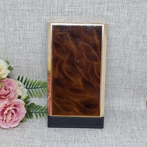 May include: A rectangular, vintage cigarette case with a gold-colored frame and a dark brown, wood-grain patterned center. The bottom is black. The case is standing upright on a light gray surface.