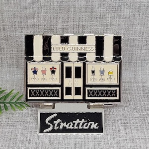 May include: A black and white compact mirror with a storefront design featuring the text "Lulu Guinness" and the word "Stratton" on a black sign.