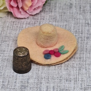 Puede incluir: Un sombrero de fieltro beige con una corona cilíndrica a juego, decorado con bayas y hojas de fieltro. Una dedal de plata vintage con un diseño floral está junto al sombrero. El fondo incluye una flor rosa y una superficie gris texturizada.