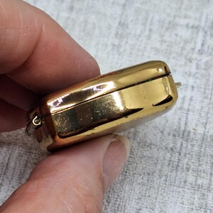 May include: A rectangular, gold-colored jewelry box with rounded edges. The box is closed, with a visible hinge and clasp. The surface reflects light, suggesting a polished finish. The box is held in a hand.