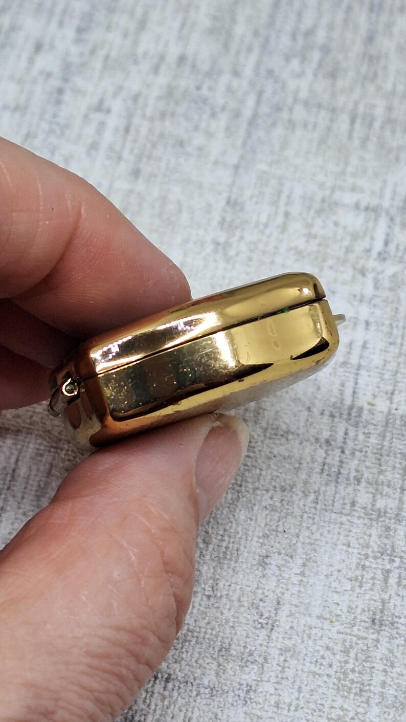 May include: A rectangular, gold-colored jewelry box with rounded edges. The box is closed, with a visible hinge and clasp. The surface reflects light, suggesting a polished finish. The box is held in a hand.