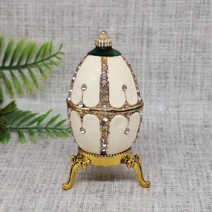 May include: An egg-shaped trinket box with a cream-colored enamel finish, accented with gold trim and sparkling rhinestones. The hinged lid is topped with a gold crown and green accents. The box rests on ornate gold-colored legs.
