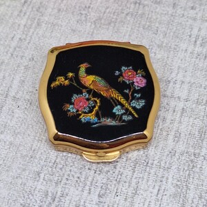 May include: A vintage compact mirror with a gold-tone frame and a black enamel top. The top features a colorful illustration of a pheasant perched on a branch with pink and red flowers. The compact is square with rounded edges.