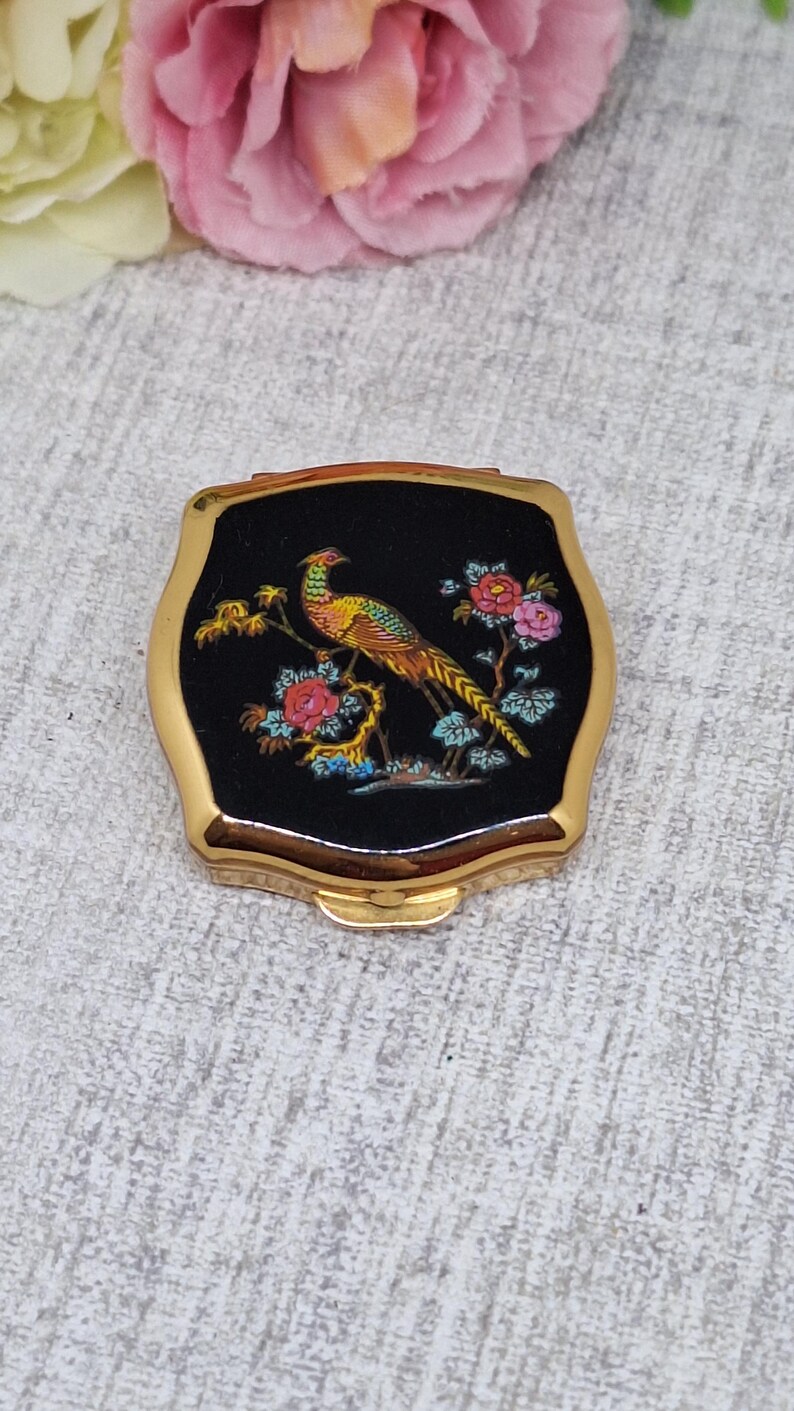 May include: A vintage compact mirror with a gold-tone frame and a black enamel top. The top features a colorful illustration of a pheasant perched on a branch with pink and red flowers. The compact is square with rounded edges.