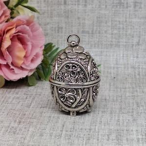 Peut inclure: Boîte à bijoux argentée en forme d'œuf avec un design floral en filigrane complexe. La boîte à charnière comporte une boucle en haut et repose sur trois petits pieds. Le travail du métal détaillé crée un aspect décoratif et orné.