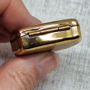 May include: A small, rectangular, gold-colored hinged box. The box has a shiny, reflective surface and appears to be made of metal. The box is held in a person's hand, with the background out of focus.
