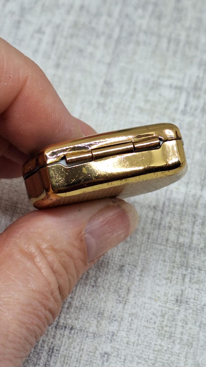 May include: A small, rectangular, gold-colored hinged box. The box has a shiny, reflective surface and appears to be made of metal. The box is held in a person's hand, with the background out of focus.
